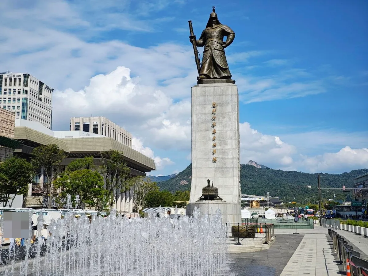 The bronze statue of Admiral Yi Sun-sin standing with a sword on a tall stone pedestal engraved with the vertical characters 忠武公李舜臣將軍像, a turtle ship model at its base and fountains spraying in the foreground, with the Sejong Center and mountains behind (faces blurred)