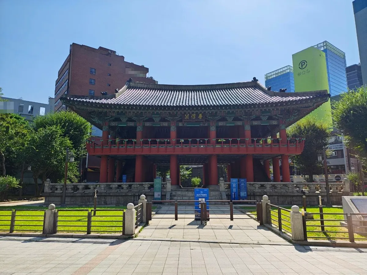 The Bosingak bell pavilion in Jongno, Seoul: a two-story traditional pavilion with red columns, painted beams and a curved tiled roof, behind a low stone fence and lawn, with modern buildings around it