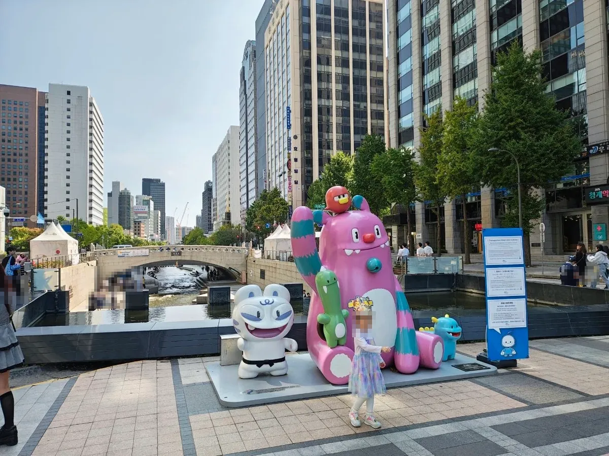The Haechi and Soul Friends character statues on a stone plaza beside Cheonggyecheon, with a stone bridge over the stream and office towers behind (faces blurred)