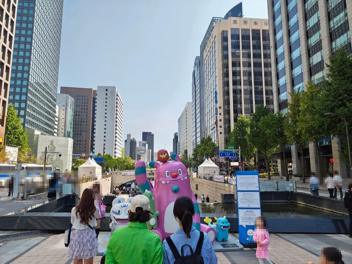 Colorful character statues at the start of Cheonggyecheon: a pink furry monster with a red bird on its head, a white tiger-like character and small green and blue creatures, surrounded by visitors taking photos (faces blurred)
