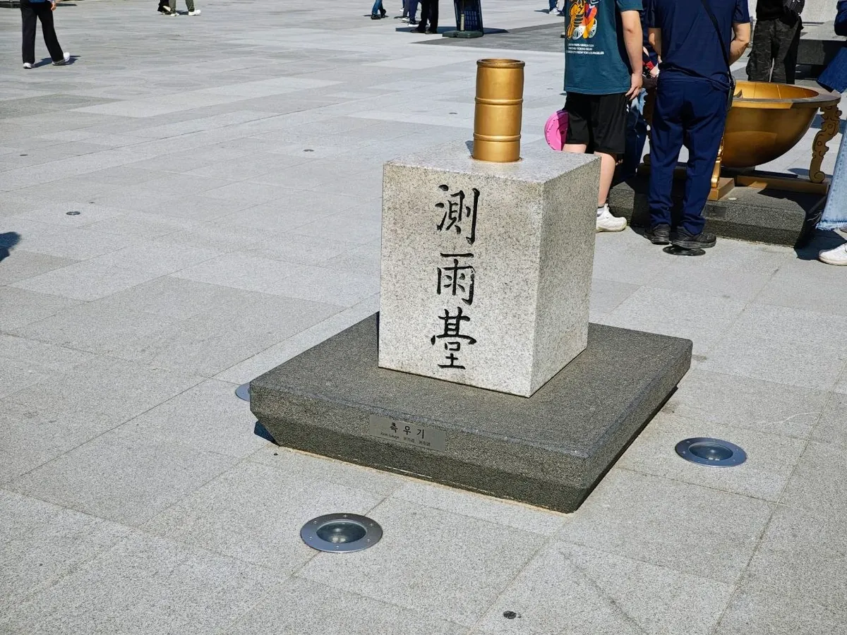 A copy of the Joseon rain gauge: a golden cylinder set into a granite block carved with the characters 測雨臺, on a dark stone base with a small plaque reading 측우기 Rain Gauge, in Gwanghwamun Square