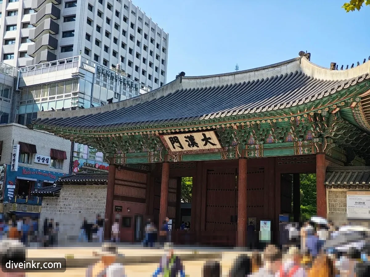 Daehanmun, the main gate of Deoksugung Palace: a wooden gate with a tiled roof, painted eaves, red pillars and doors and a name board reading ๅคงๆผข้, standing between office buildings, with visitors in front (faces blurred)