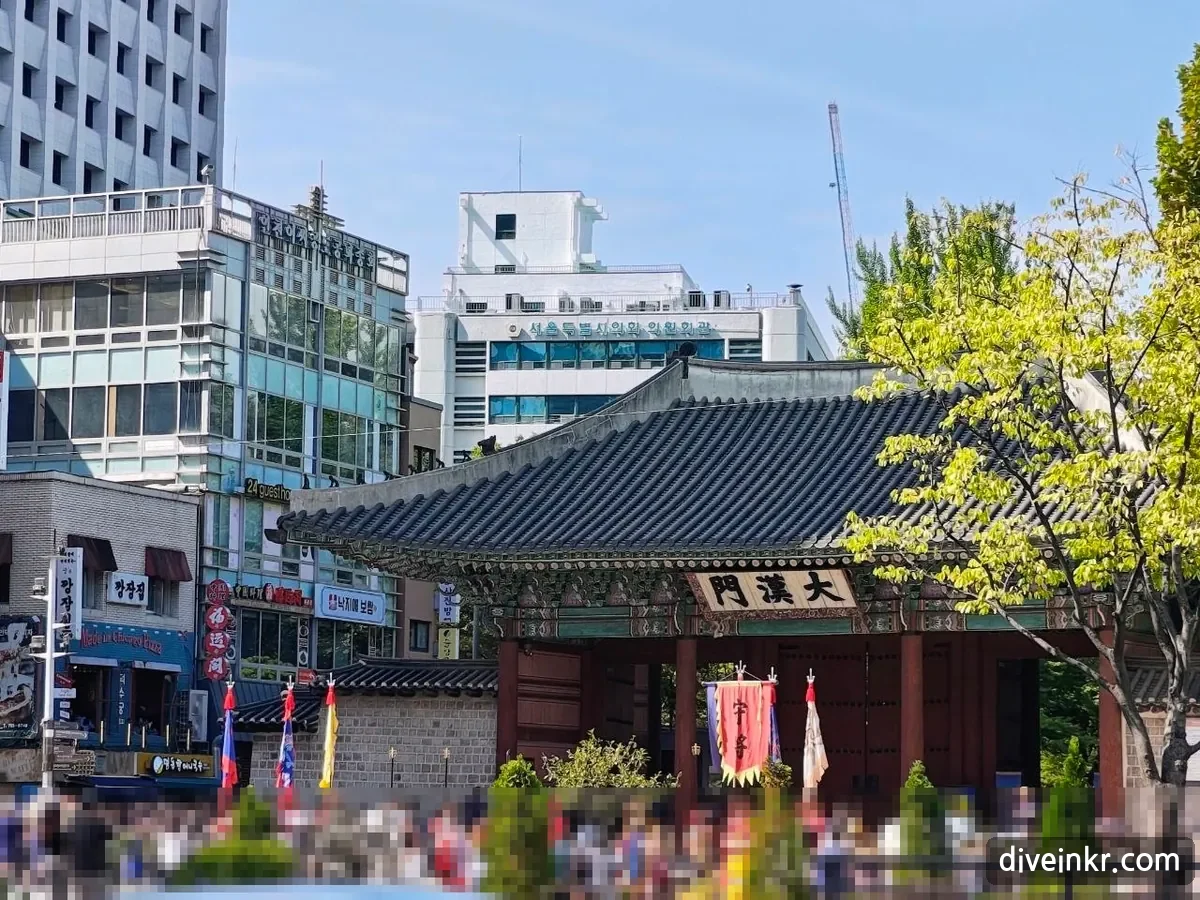 Tall red, blue, yellow and white flags of the royal guard ceremony raised in front of Daehanmun gate, seen from a distance over a crowd of spectators (faces blurred), with office buildings behind the gate