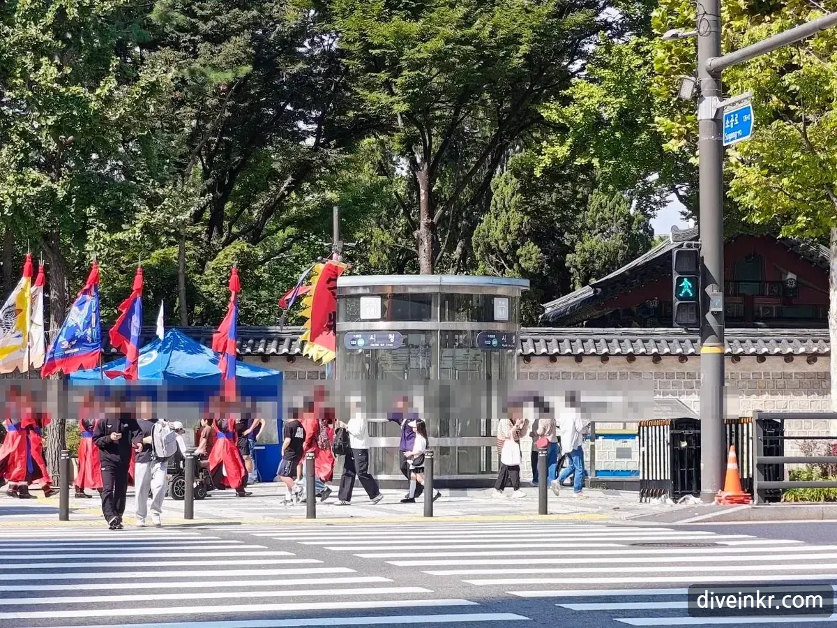 Guards in red Joseon-era uniforms carrying tall red, blue and yellow flags on the sidewalk beside Deoksugung's stone wall, next to a glass elevator entrance marked ์์ฒญ with the station numbers 132 and 201, seen across a crosswalk; a blue street sign on the right reads ์๊ณต๋ก Sogong-ro (faces blurred)