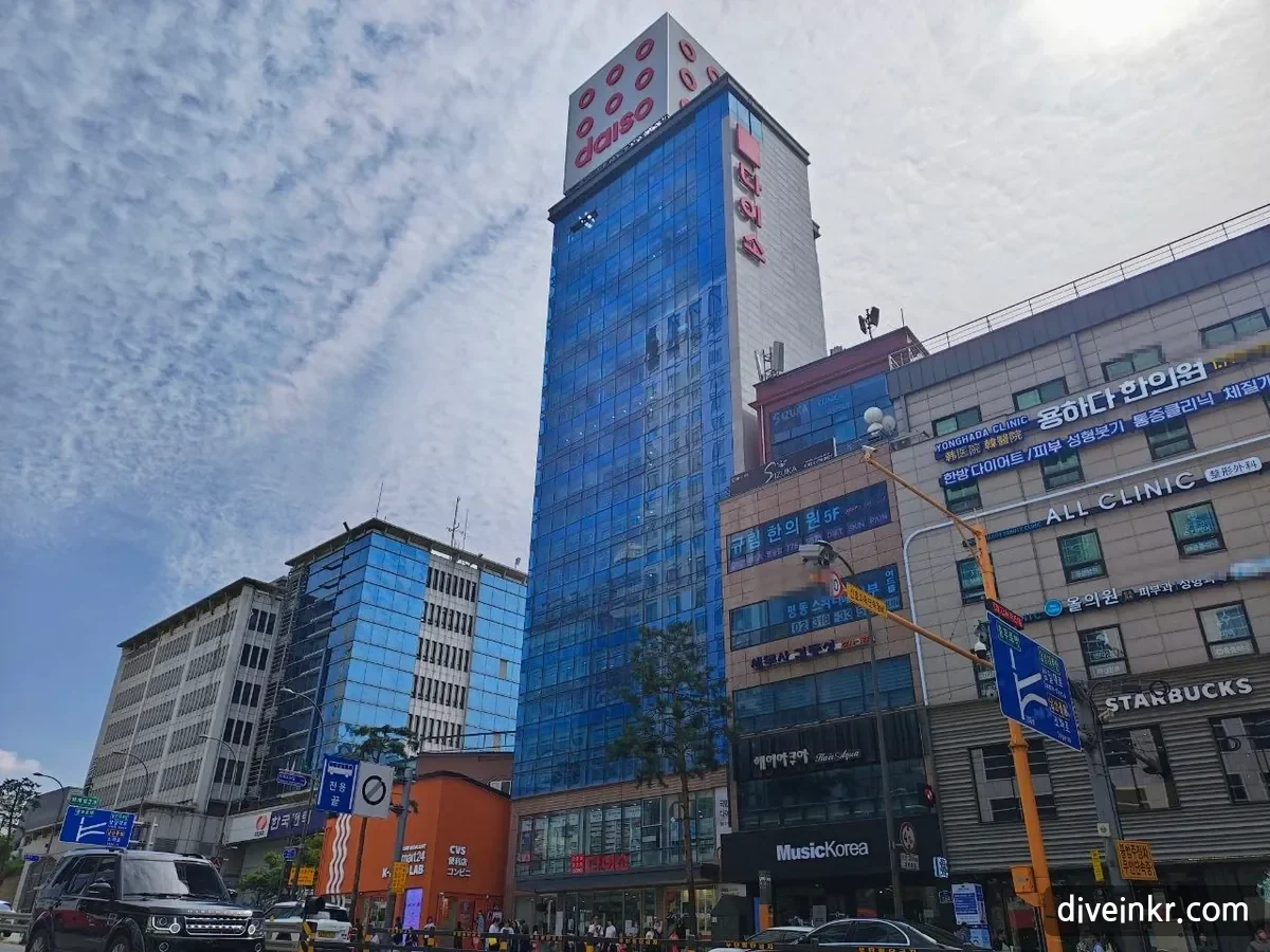 The Daiso Myeongdong Station store: a tall narrow blue-glass building with a red daiso logo and the vertical Korean sign 다이소 at the top, next to buildings with signs for a Korean medicine clinic, other clinics and a Starbucks, under a sky streaked with clouds