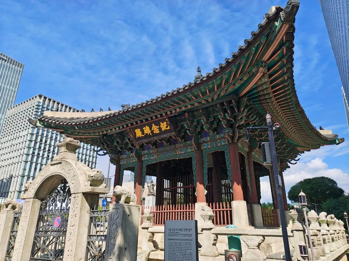 The pavilion for the monument to King Gojong's 40th year on the throne: a richly painted wooden pavilion with sharply upturned eaves and a name board reading 紀念碑殿, behind a stone gate topped with stone animals and an information panel, next to glass office towers