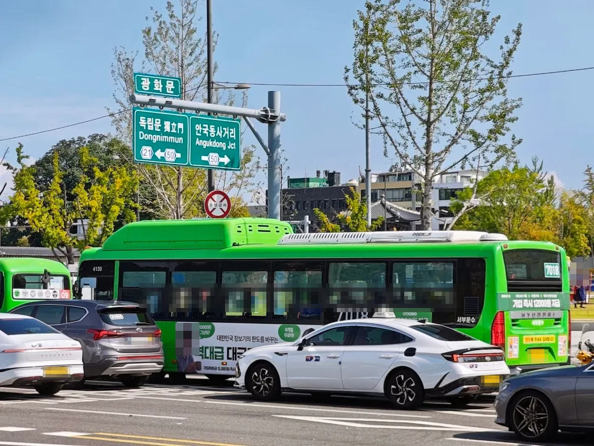 A green road sign at the Gwanghwamun intersection pointing left to 독립문 Dongnimmun and right to 안국동사거리 Angukdong Jct, above a green Seoul bus (route 7018), a white taxi and cars waiting in traffic (license plates and an ad model's face blurred)