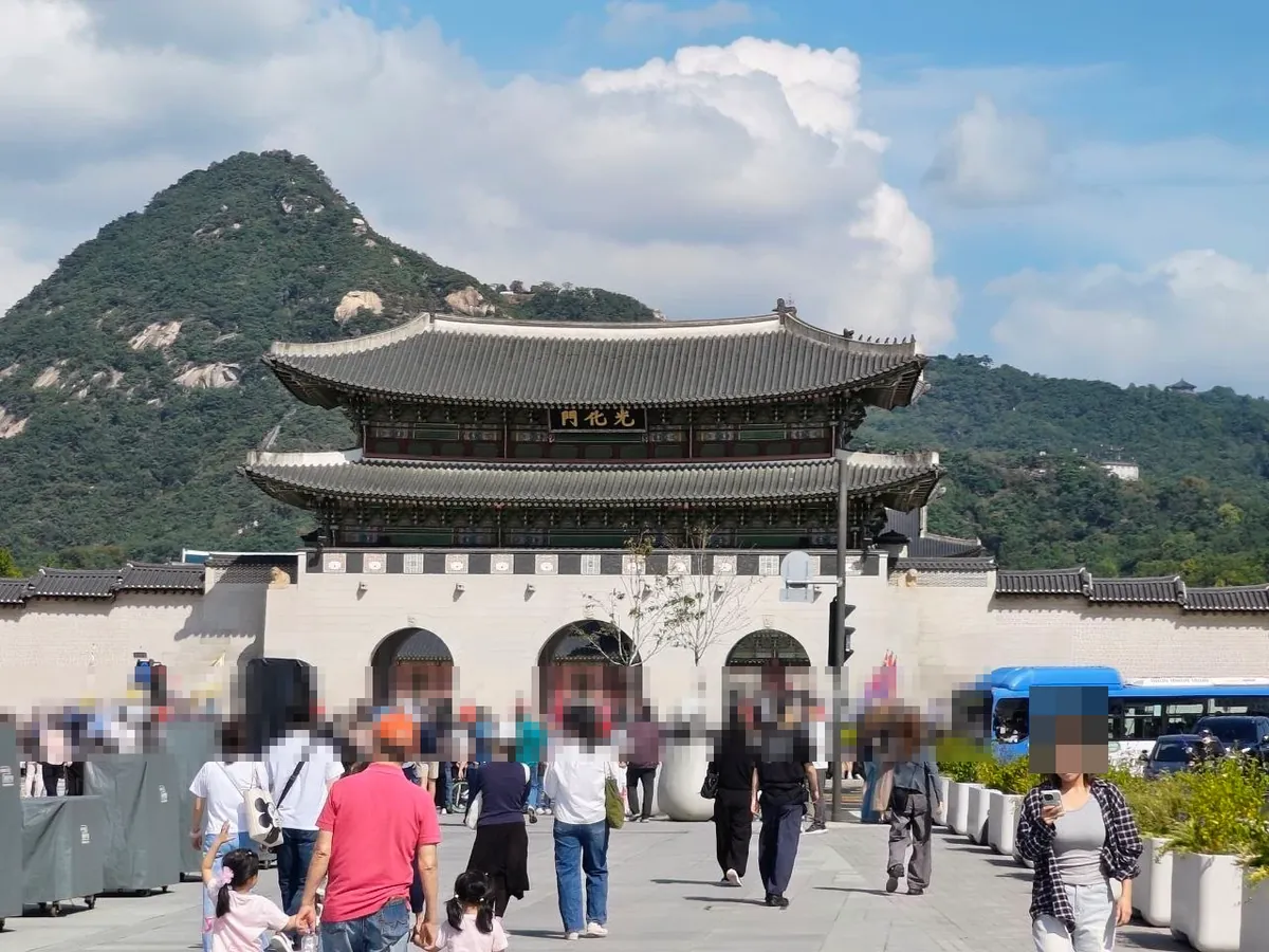 Gwanghwamun, the main gate of Gyeongbokgung Palace: a two-story wooden gate tower with a curved tiled roof and a black name board with gold characters 光化門, on a stone base with three arched openings, crowds of visitors in front and Bugaksan behind (faces blurred)