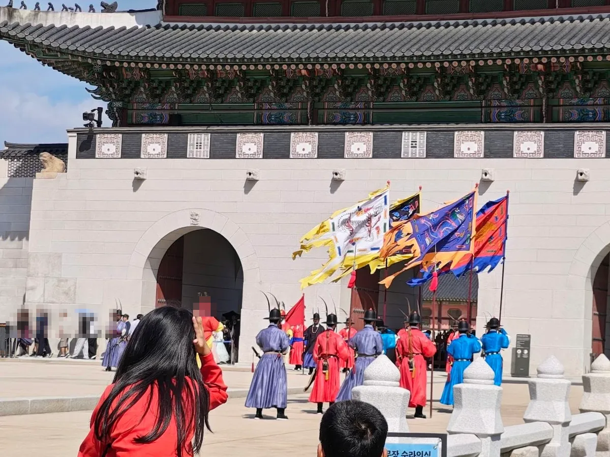 Close-up of the guards: men in red, blue and purple robes with black feathered hats and bows on their backs stand in formation in front of the stone base of Gwanghwamun, holding large painted flags, one showing a dragon, as spectators watch in the foreground; a blue sign at the bottom edge reads 순라의식 (faces blurred)