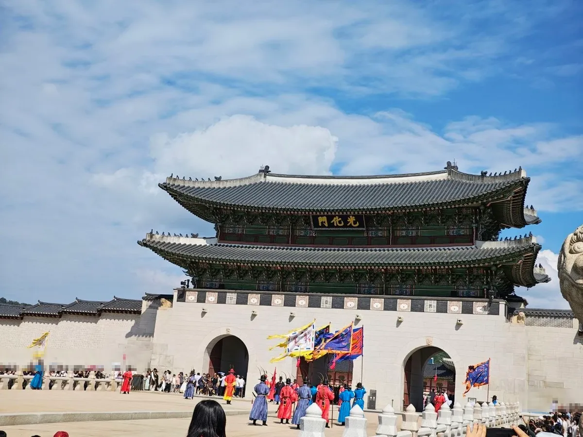 The royal guard ceremony in front of Gwanghwamun: guards in red, blue and purple Joseon-era uniforms stand in formation in front of the arched gateways, holding tall colorful flags, while crowds watch from behind stone railings (spectators' faces blurred)
