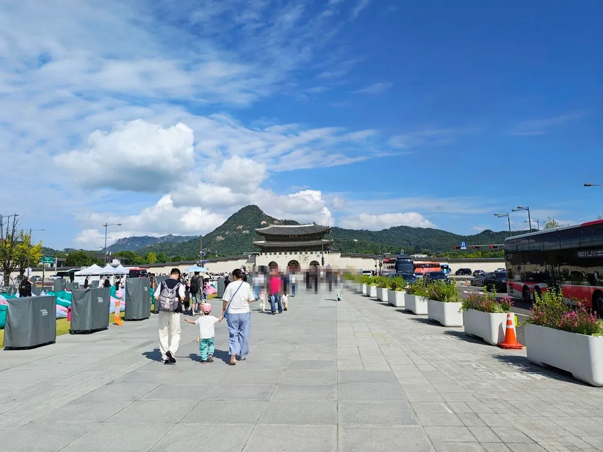 The wide stone walkway of Gwanghwamun Square leading north to Gwanghwamun gate, with Bugaksan mountain behind it under a blue sky, families walking, planters along the road on the right and a red express bus passing