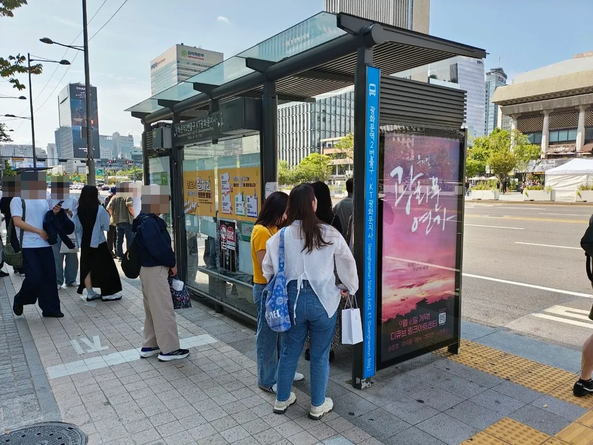 A bus shelter on the road beside Gwanghwamun Square with a blue sign reading 광화문역2번출구·KT광화문지사 and the English Gwanghwamun Station Exit 2 · KT (Gwanghwamun Bldg.), people waiting and a musical poster on the side panel (faces blurred)
