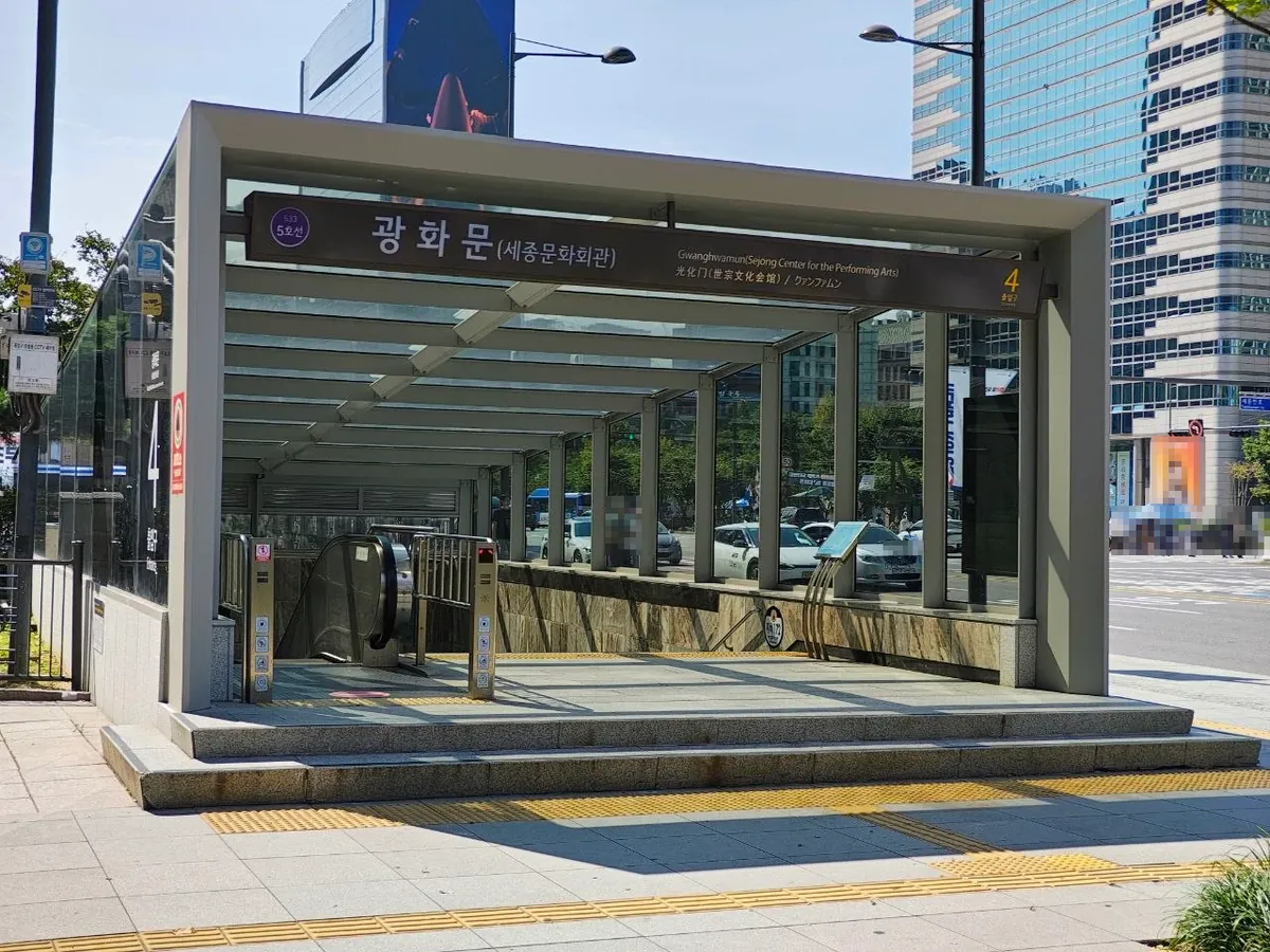 The glass-roofed entrance to exit 4 of Gwanghwamun Station on subway Line 5, with a sign reading 광화문(세종문화회관) Gwanghwamun (Sejong Center for the Performing Arts) and an escalator going down (faces blurred)