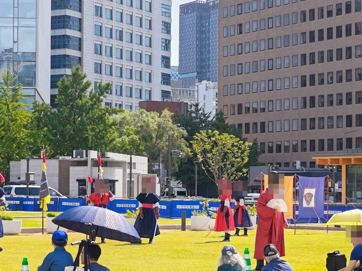 Performers in Joseon-era costumes — red and navy military uniforms with feathered hats, and a man in a red official's robe holding a fan — performing on the lawn beside Gwanghwamun Square, with banners and an audience seated on the grass (faces blurred)