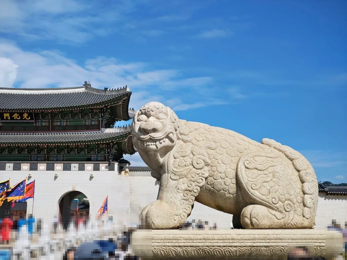 A large stone haechi statue with curly carved mane, bulging eyes and a toothy grin, crouching on a stone plinth in front of Gwanghwamun gate, with guards in colorful uniforms and flags by the gate and visitors below (faces blurred)