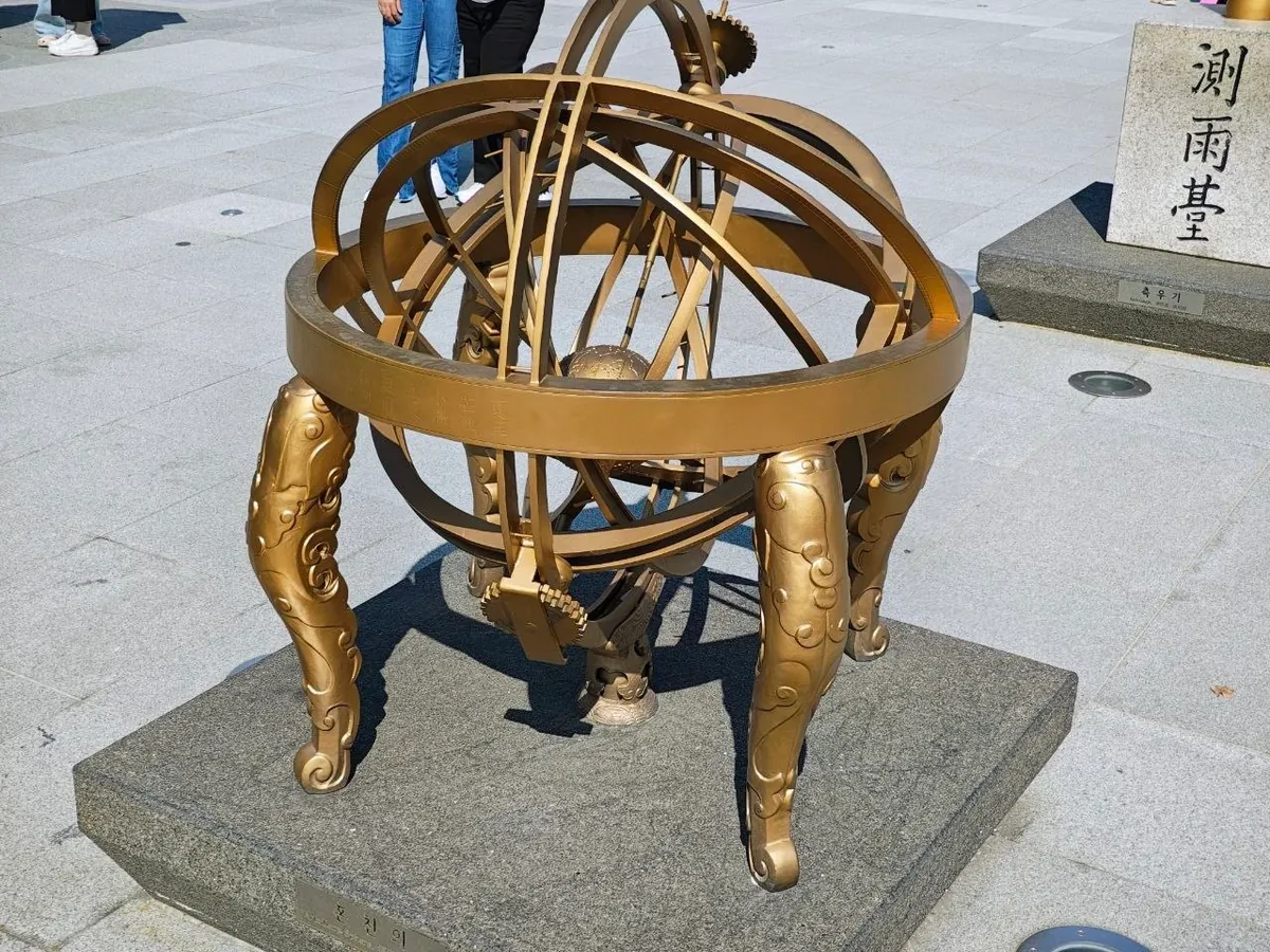 A golden copy of a Joseon armillary sphere: interlocking rings with a small globe in the center, set on four curved legs decorated with cloud patterns, on a stone base in Gwanghwamun Square