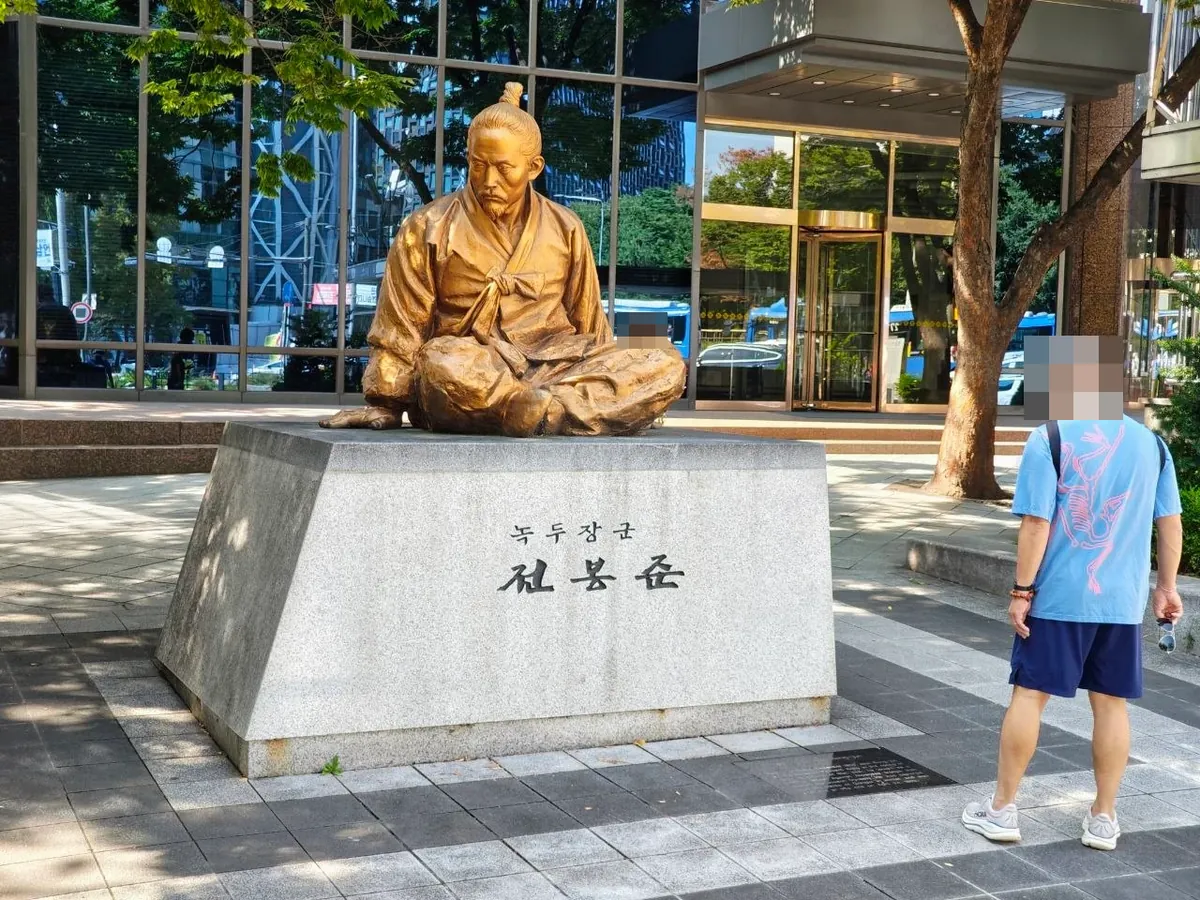 A bronze statue of Jeon Bong-jun seated cross-legged in traditional clothes on a granite base engraved 녹두장군 전봉준, outside a glass building near Jonggak Station, with a passerby standing beside it (face blurred)