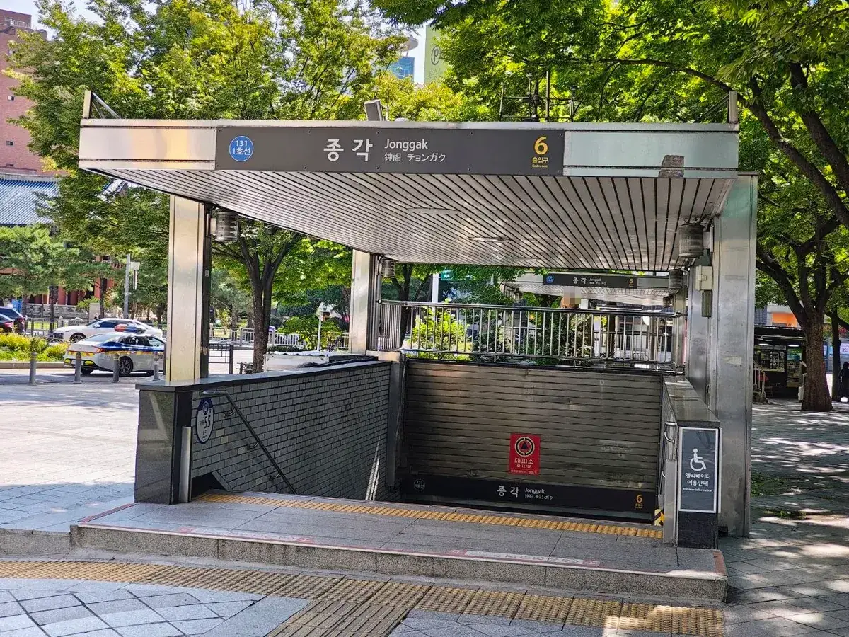 The covered entrance to exit 6 of Jonggak Station on Seoul subway Line 1, with a sign reading 종각 Jonggak 钟阁 チョンガク and the number 6, stairs leading down past a red 대피소 SHELTER sign, and trees and traffic behind