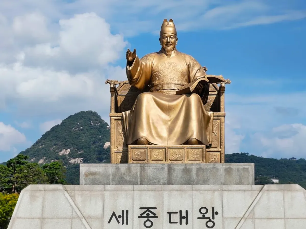 The golden bronze statue of King Sejong seated on a throne, holding an open book in his left hand and raising his right hand, on a stone pedestal engraved 세종대왕 in Hangul, with a green mountain behind