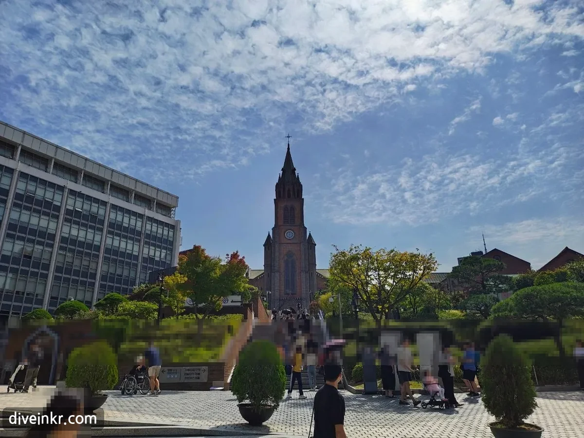 Myeongdong Cathedral, a red-brick Gothic church with a tall pointed bell tower and a clock, at the top of a wide stone staircase lined with trees, with visitors climbing the steps and walking on the plaza below under a blue sky with clouds (faces blurred)