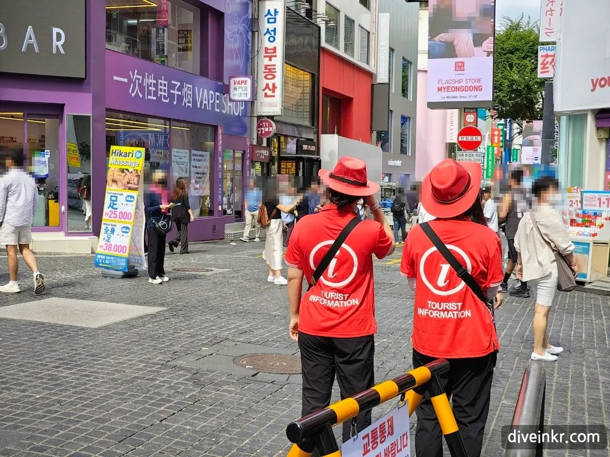 Two roaming tourist guides seen from behind in Myeongdong, wearing wide-brimmed red hats and red shirts printed with a white i and TOURIST INFORMATION, standing behind a yellow-and-black barrier with a sign reading 교통통제 and looking out over a busy shopping street with a no-entry sign in the distance (bystanders' faces blurred)