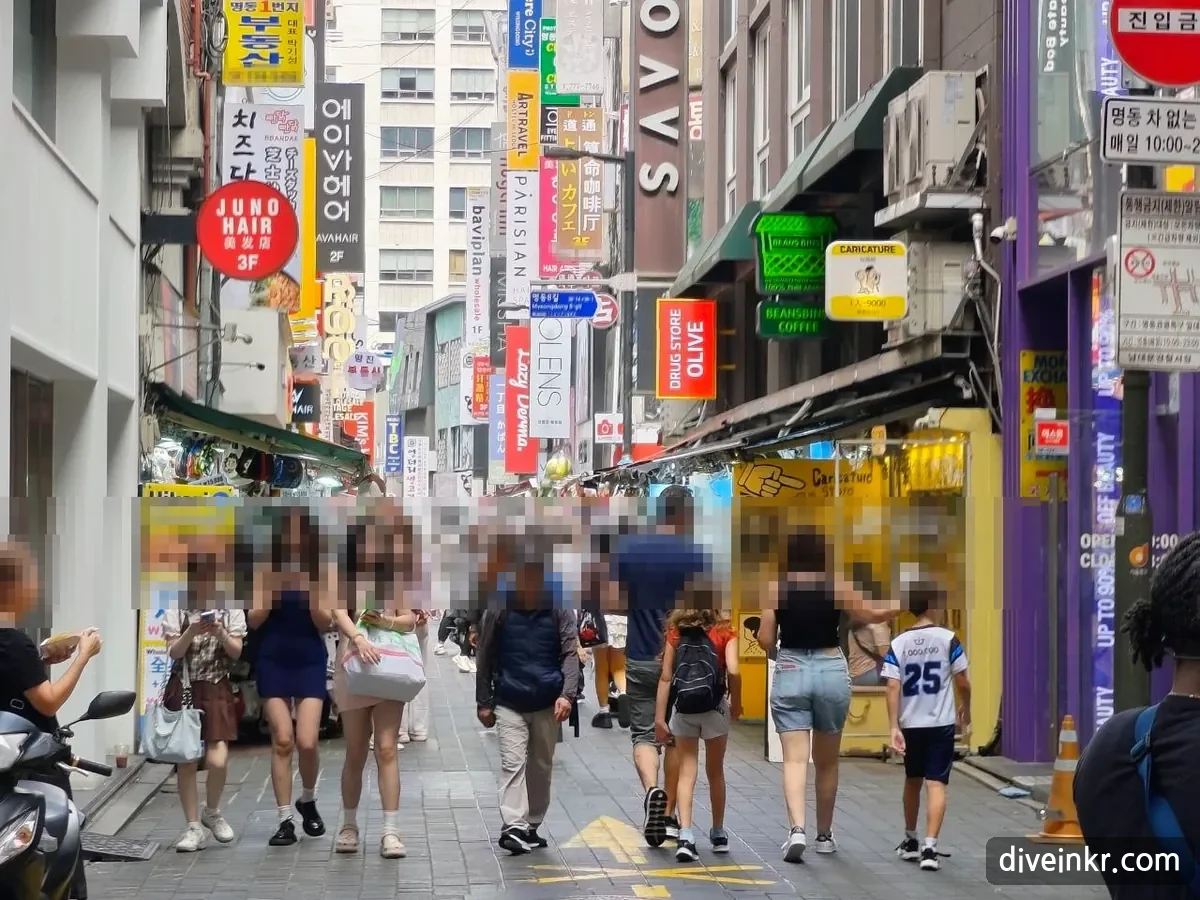 A narrow Myeongdong side street crowded with shoppers under dozens of vertical signs in Korean, English, Chinese and Japanese, including JUNO HAIR 3F, DRUG STORE OLIVE, a caricature shop and a small blue street sign reading 명동8길 Myeongdong 8-gil; on the right, a red no-entry sign reading 진입금지 above a sign reading 명동 차 없는 거리 매일 10:00~23:00 (faces blurred)