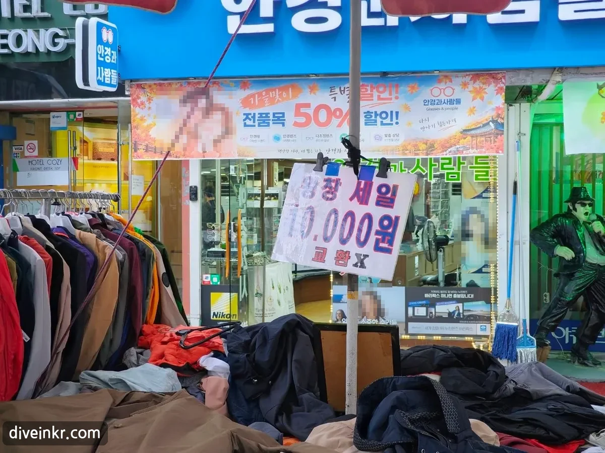 A handwritten sign clipped to a parasol pole above a pile of jackets, reading ์์ฐฝ ์ธ์ผ 10.000์ ๊ตํ X. Behind it is an optician's shop with a blue sign and an autumn sale banner reading ๊ฐ์๋ง์ด ํน๋ณํ ์ธ! ์ ํ๋ชฉ 50% ํ ์ธ! (faces blurred)