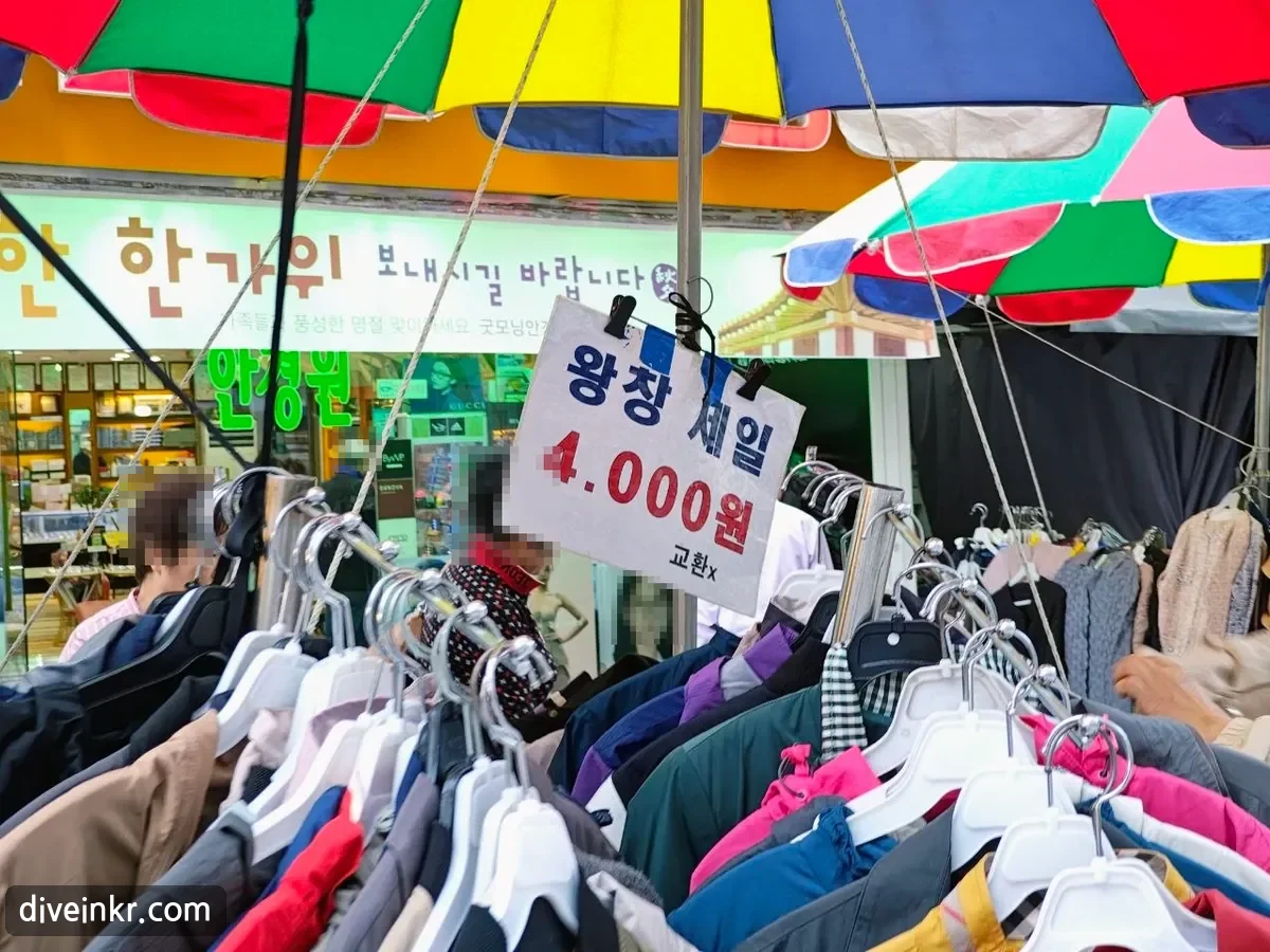 A handwritten sign reading ์์ฐฝ ์ธ์ผ 4.000์ ๊ตํx clipped above racks of jackets under colorful parasols, with a shop sign behind reading ํ๊ฐ์ ๋ณด๋ด์๊ธธ ๋ฐ๋๋๋ค and a green optician's sign (faces blurred)