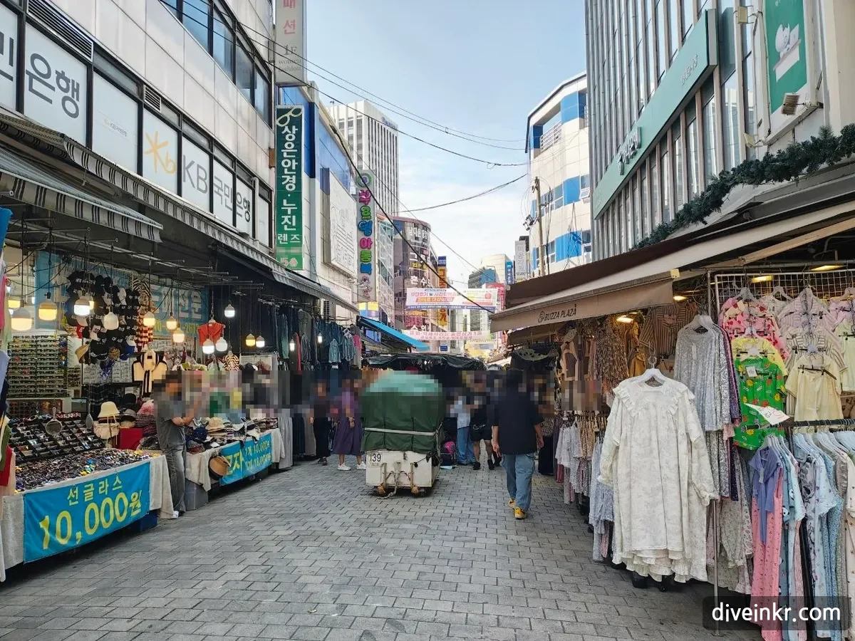 A street in Namdaemun Market lined with stalls selling sunglasses and hats, with a blue banner reading ์ ๊ธ๋ผ์ค 10,000์, racks of dresses on the right, a covered cart in the middle of the lane, a KB Kookmin Bank sign on the building at left and shoppers walking (faces blurred)