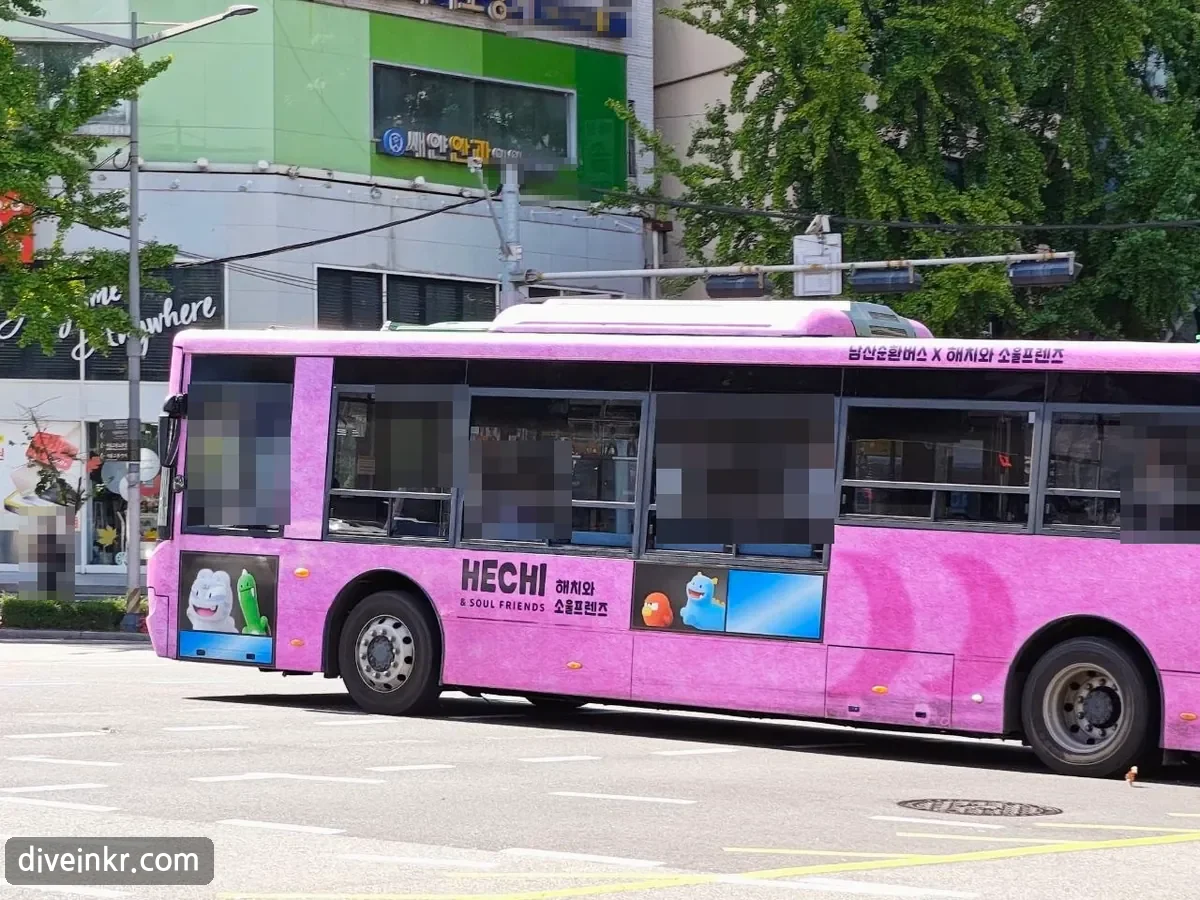 A pink 남산순환버스 (Namsan loop bus) wrapped with the 해치와 소울프렌즈 characters, driving past shops (passengers' faces blurred)
