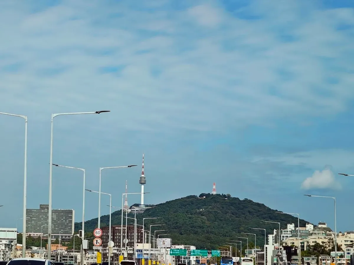N Seoul Tower standing on the green hill of Namsan under a blue sky with light clouds, seen from a road in Seoul lined with street lamps and road signs