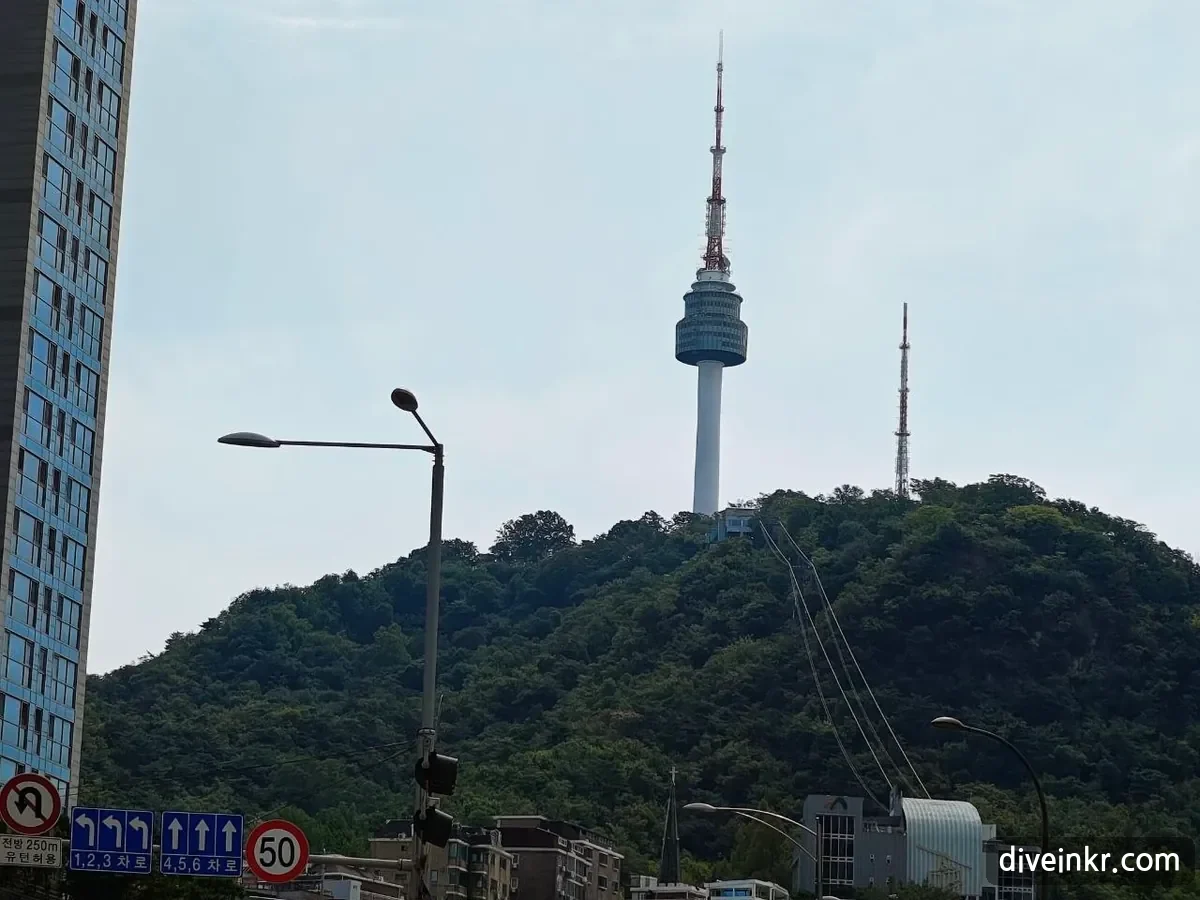 N Seoul Tower on the wooded summit of Namsan seen from below, with a second transmission mast to the right and cables running down the hillside, above a no-U-turn sign with a plate reading 전방 250m 유턴허용, blue lane signs reading 1,2,3 차로 and 4,5,6 차로, and a 50 km/h speed-limit sign