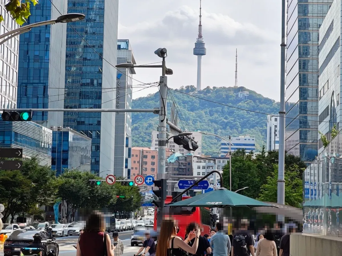 N Seoul Tower on the green hill of Namsan seen past traffic lights and glass towers on a busy downtown street near Cheonggyecheon, with a 청계천로 street-name sign, a red tour bus and pedestrians (faces and license plates blurred)