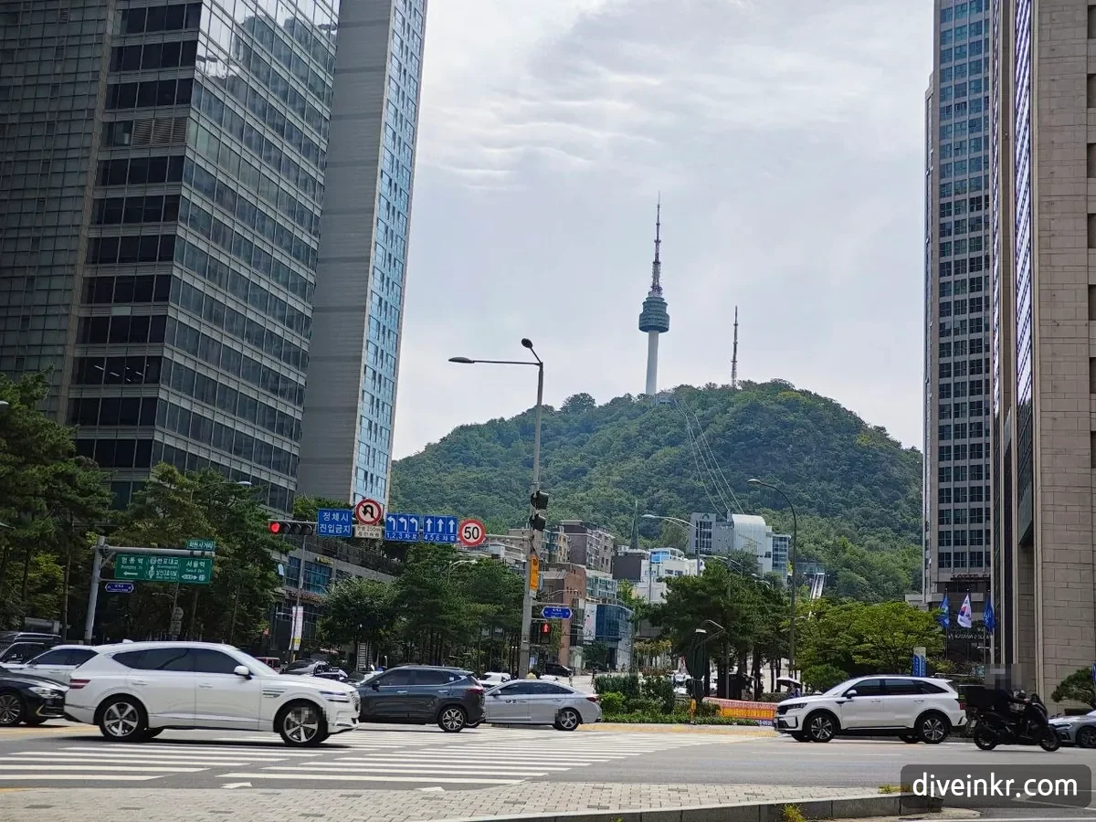 A wide downtown intersection with cars crossing, office towers on both sides, and Namsan with N Seoul Tower rising in the gap between the buildings; a green direction sign at left, under a smaller sign reading 회현사거리, points left to 명동역 and right to 서울역 (license plates blurred)