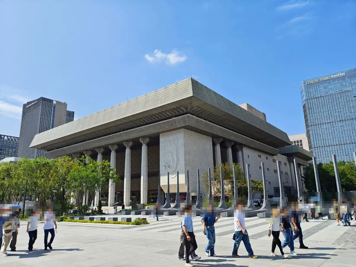 The Sejong Center for the Performing Arts on the west side of Gwanghwamun Square: a large stone building with a wide overhanging roof and tall columns, with visitors walking across the plaza in front (faces blurred)