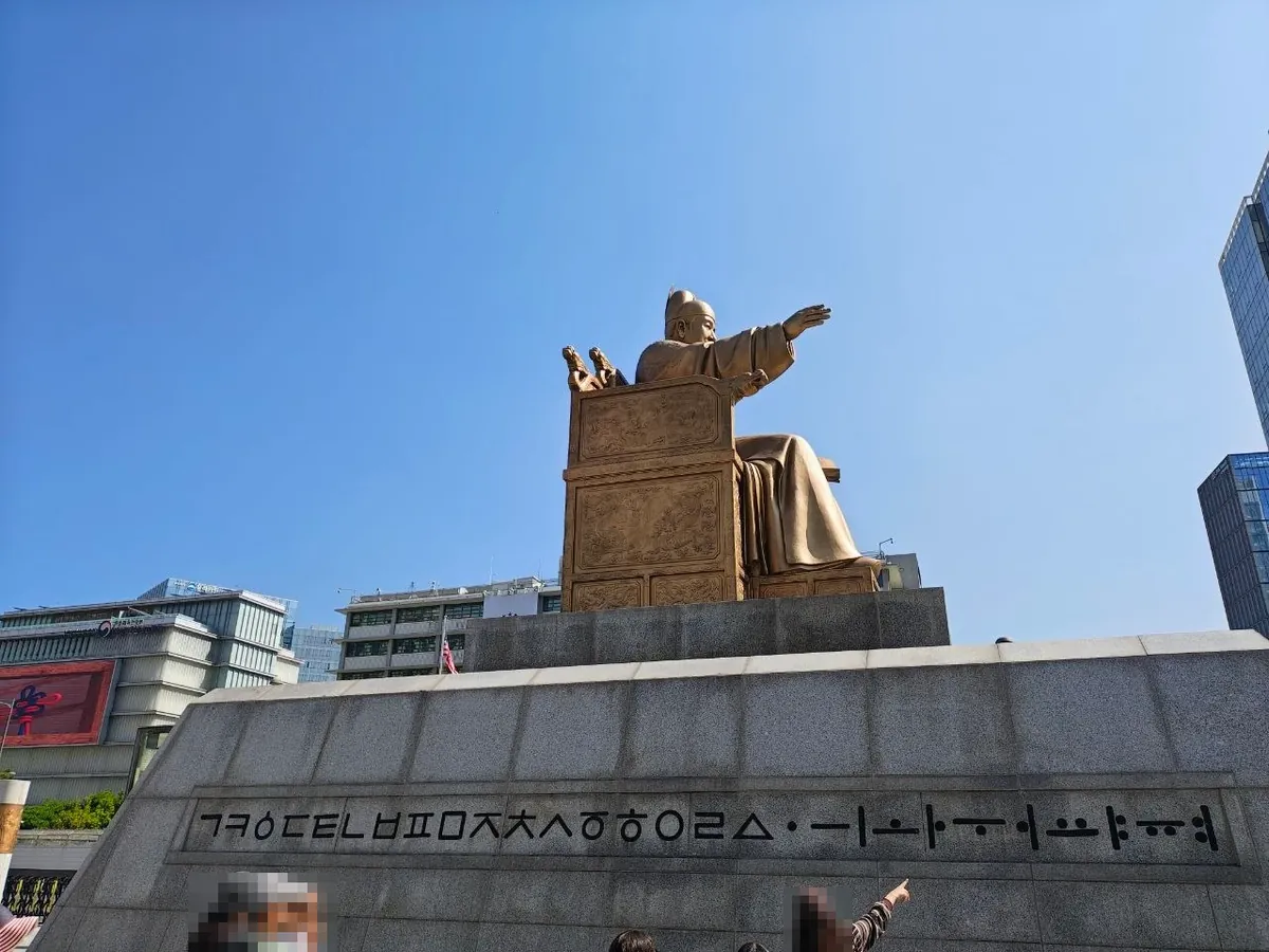 The King Sejong statue seen from the side and slightly behind, seated on a carved throne with his right arm stretched forward, above a long granite pedestal carved with a row of Hangul letters: ㄱㅋㆁㄷㅌㄴㅂㅍㅁㅈㅊㅅㆆㅎㅇㄹㅿ followed by ·ㅡㅣㅗㅏㅜㅓㅛㅑㅠㅕ, while visitors point at the letters (faces blurred)