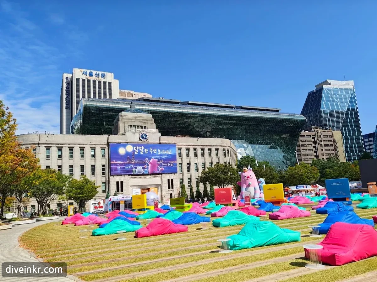 Seoul City Hall: a stone 1920s building with a clock in front of a modern glass building whose curved facade leans forward like a wave. A large screen on the old building shows the message ๋ฌ๋ฌํ ์ถ์ ๋ณด๋ด์ธ์ with a pink cartoon character and a full moon. In front, the lawn of Seoul Plaza is covered with colorful bean-bag cushions, and a large pink inflatable character stands beside a smaller blue one