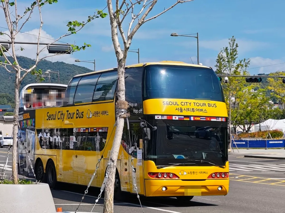 A yellow double-decker tour bus near Gwanghwamun with SEOUL CITY TOUR BUS and 서울시티투어버스 above the windshield, a row of national flags, Hop on Hop off on the side window and the 노랑풍선 시티버스 name on the front (license plate and passengers blurred)