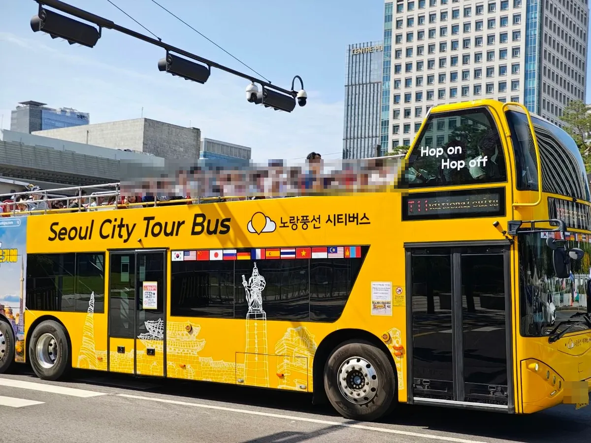 A yellow open-top double-decker bus reading Seoul City Tour Bus and 노랑풍선 시티버스, with Hop on Hop off on the front window, flags of many countries along the side and passengers on the upper deck (faces blurred), passing office buildings near Gwanghwamun
