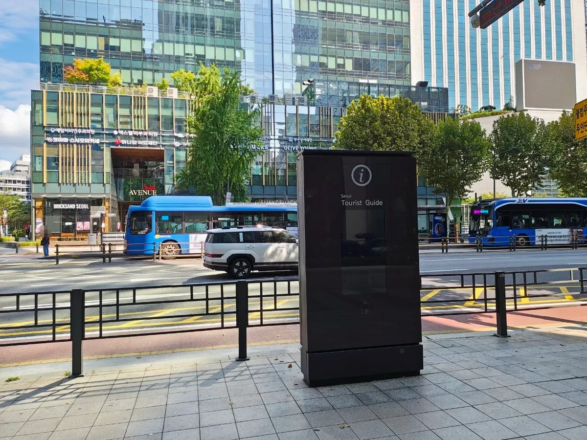 A tall black street kiosk with an i symbol and the words Seoul Tourist Guide on a city sidewalk in central Seoul, with blue buses and glass office buildings across the road