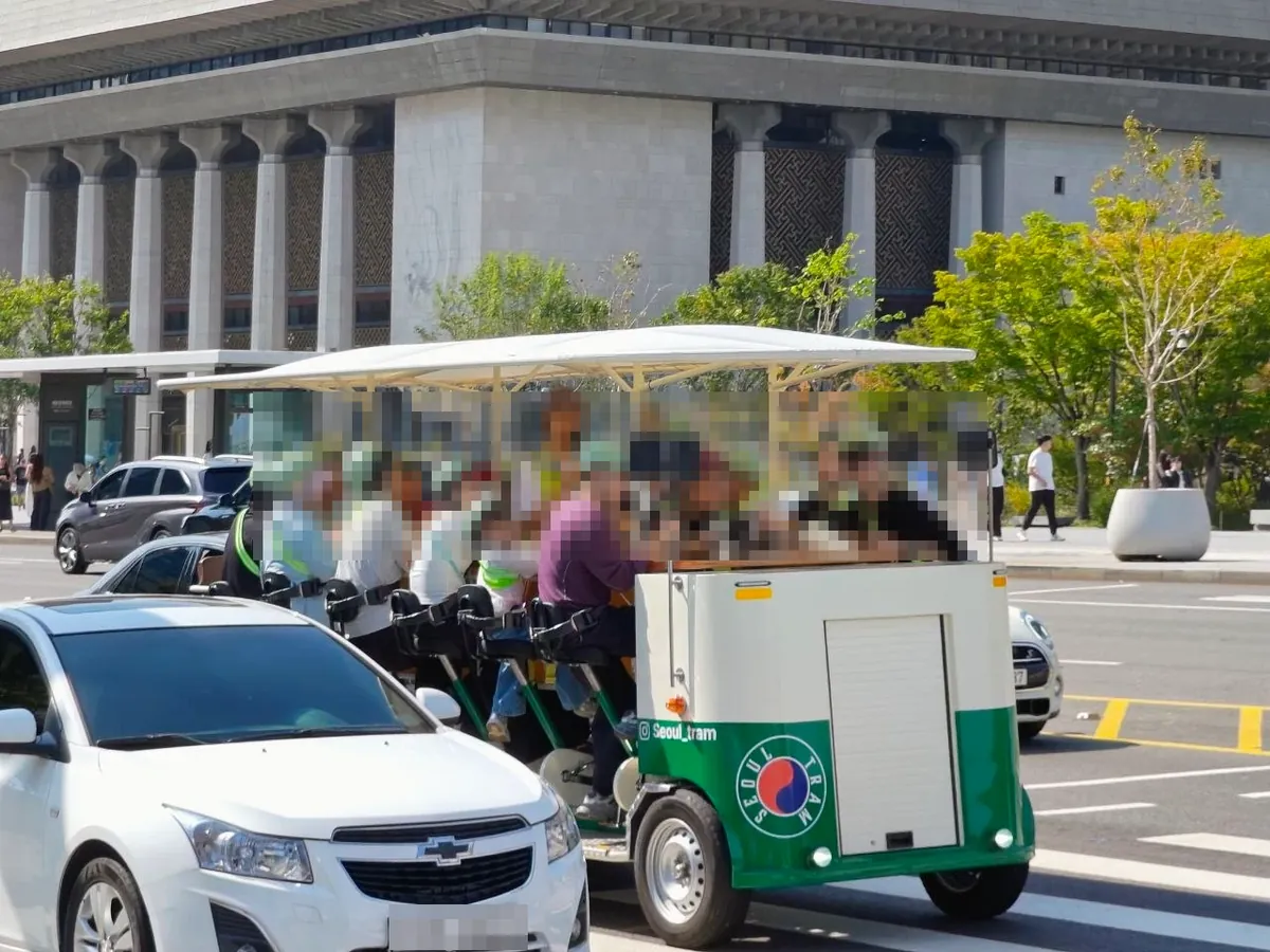 A green-and-white Seoul Tram pedal vehicle with a white canopy, carrying a group of riders who pedal together, on a road in front of the Sejong Center (faces and license plate blurred)