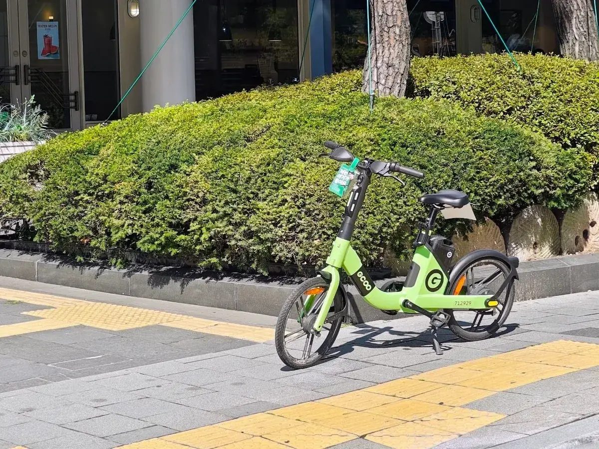 A green shared electric bicycle from a private company parked on a paved sidewalk next to a trimmed hedge in Seoul, with a yellow tactile paving strip in the foreground