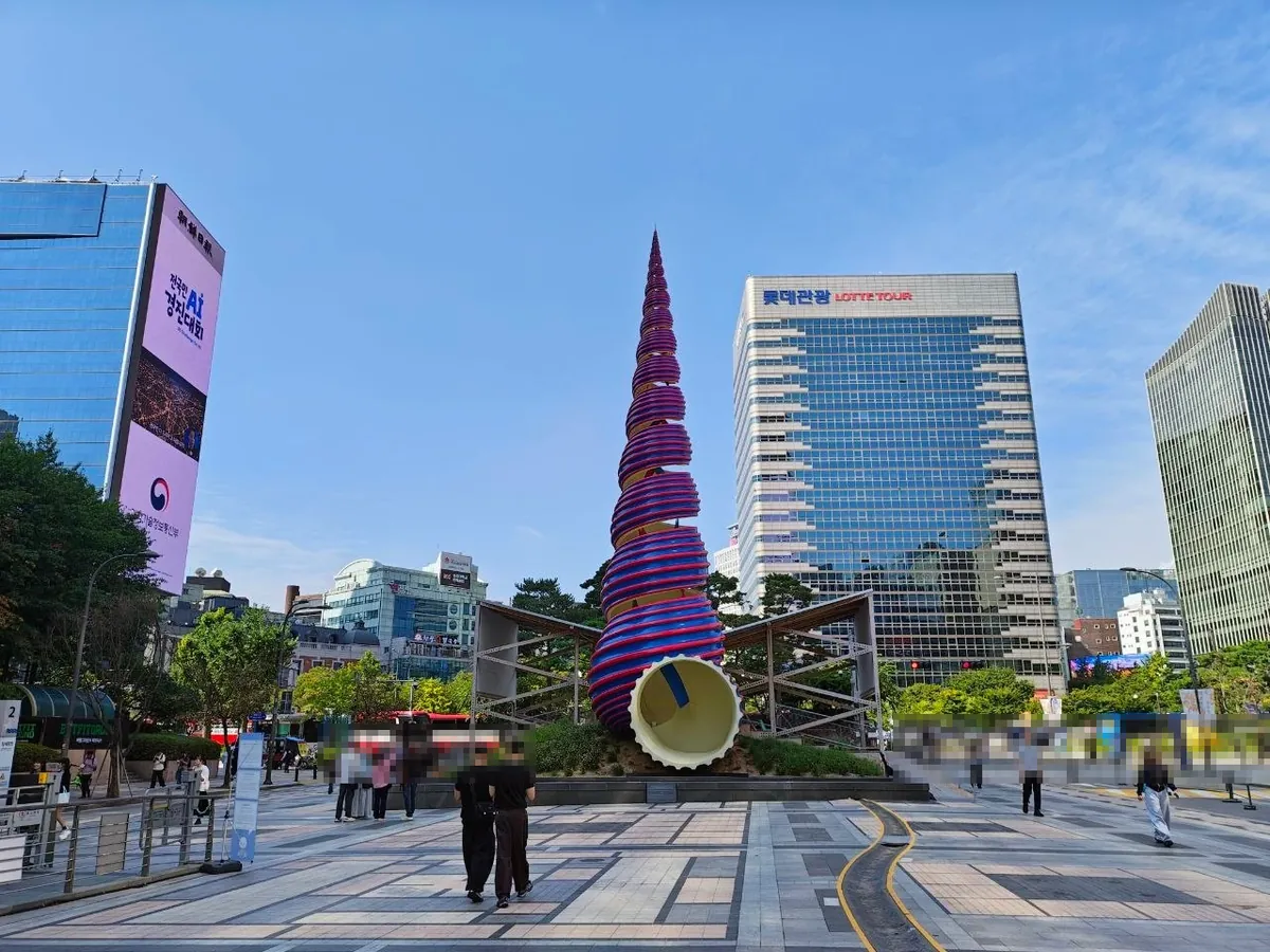 The Spring sculpture at Cheonggye Plaza: a tall spiral cone wrapped in red and blue ribbons, about 20 meters high, with a white opening at the base where the stream begins, among office towers (faces blurred)