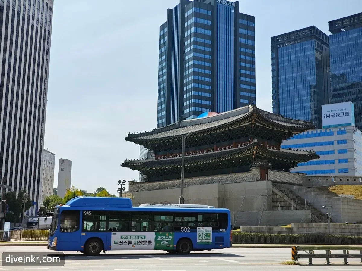 Sungnyemun on its stone base beneath tall office towers, with steps leading up beside the gate and a blue Seoul city bus, route 502, passing on the road in front