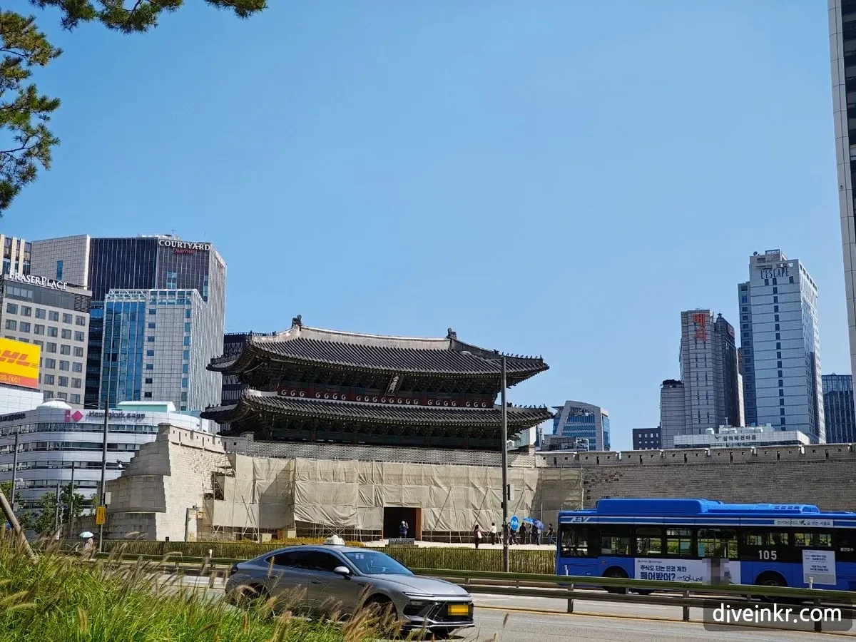 Sungnyemun, a two-story wooden gate on a massive stone base partly wrapped in beige construction screens, with a stretch of old city wall to the right and glass office towers behind, seen across a road with a blue electric city bus and a gray car passing in front and pampas grass in the foreground