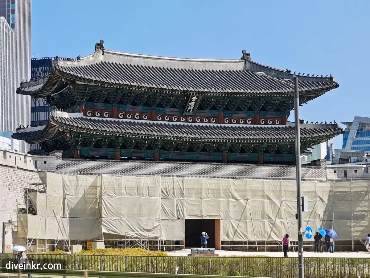 A close view of Sungnyemun's two-tiered tiled roof and painted eaves, with a vertical name board reading ๅด็ฆฎ้ hanging between the two roofs; the stone base below is wrapped in beige construction screens with an opening left at the central passage, and a few visitors stand in front