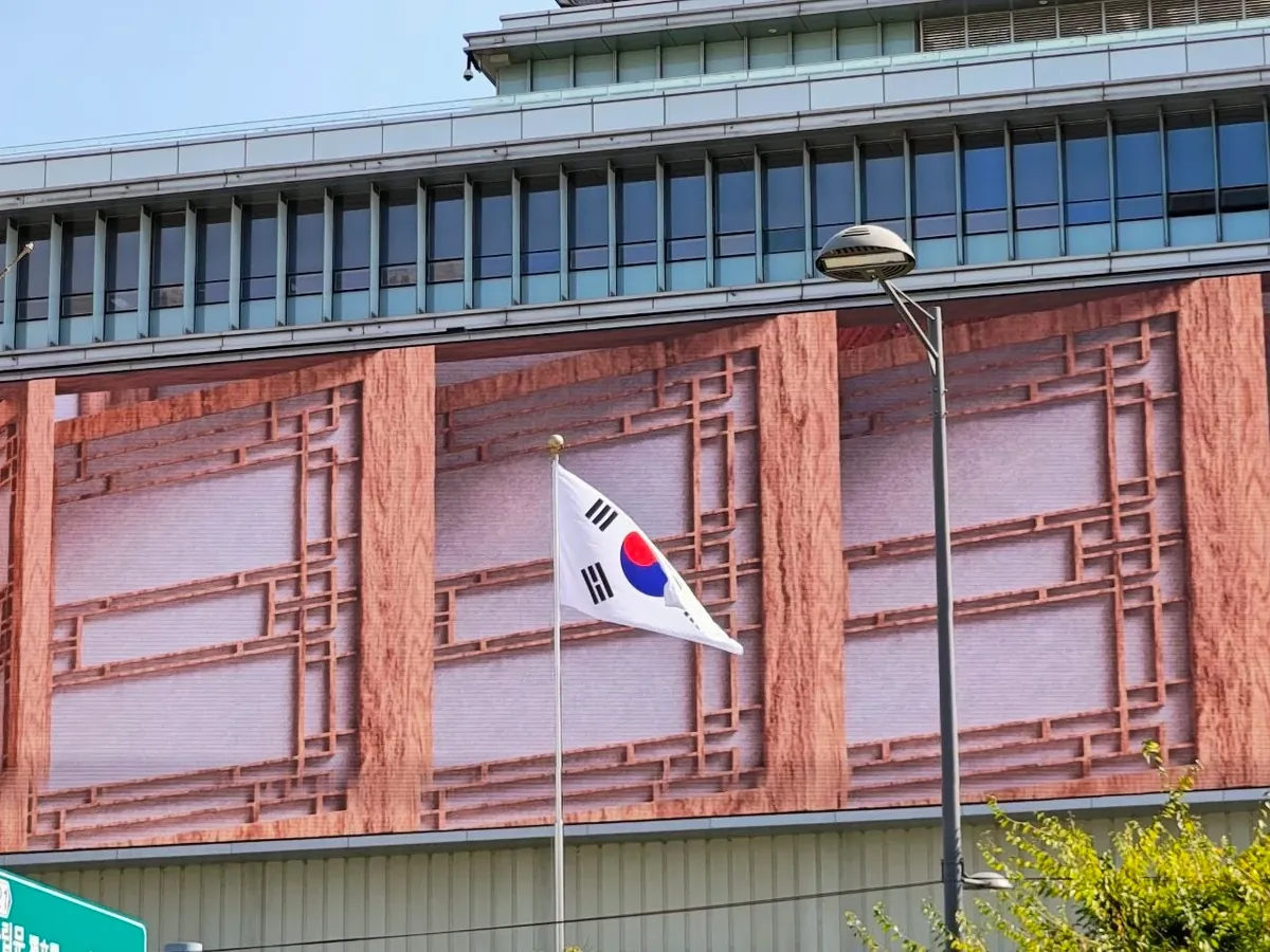 The Korean national flag, Taegeukgi, flying from a pole in front of a building near the Seoul Museum of History, whose facade shows a large image of a traditional wooden lattice window