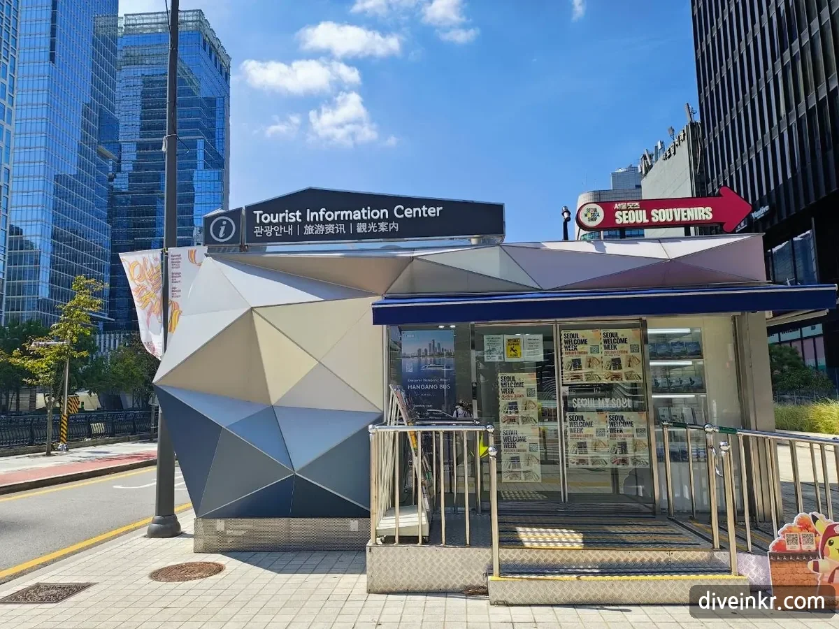 A small Tourist Information Center booth with an angular silver facade and a sign reading 관광안내 in Korean, Chinese and Japanese, with Seoul Welcome Week posters in the windows and a red arrow sign reading SEOUL SOUVENIRS beside it, on a sidewalk beside Cheonggyecheon stream