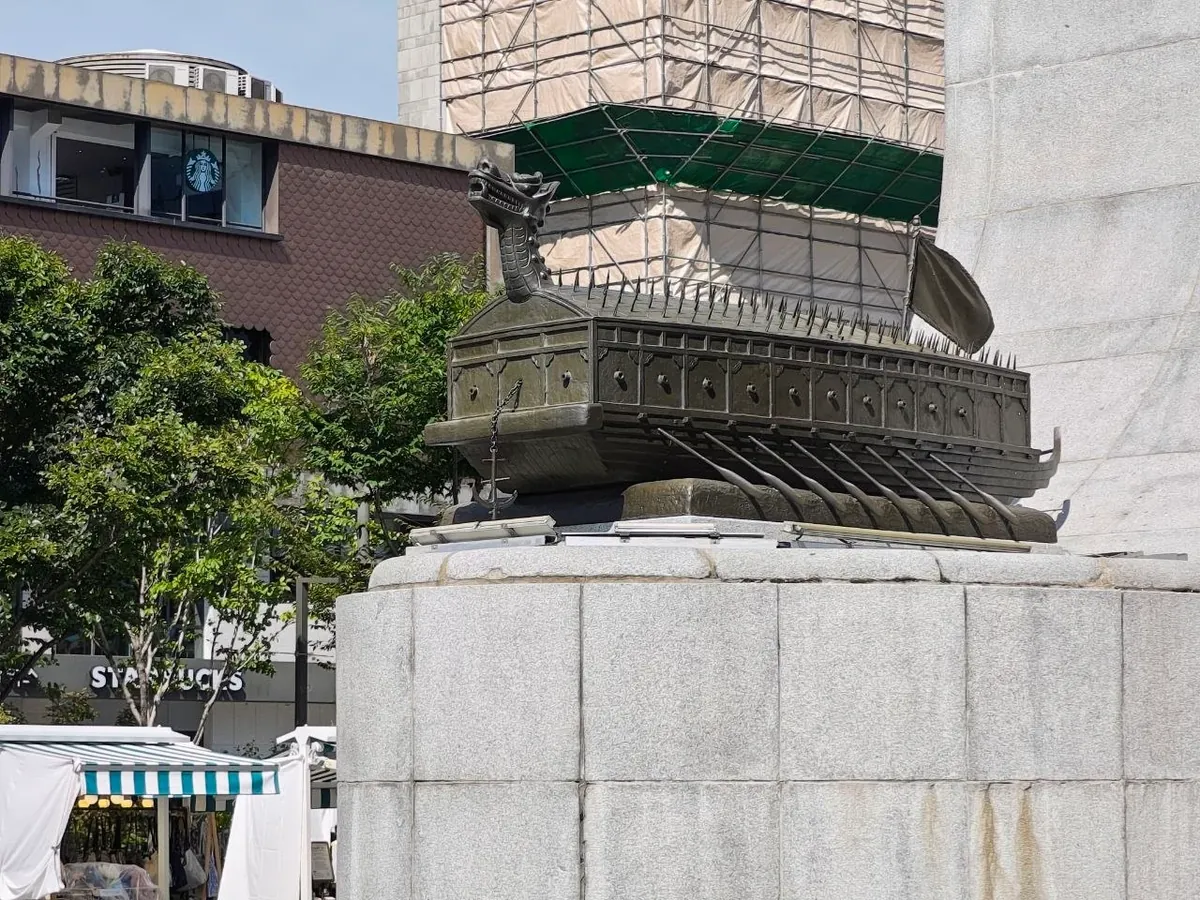 A bronze model of a turtle ship (geobukseon) with a dragon head, spiked roof and oars, sitting on the granite base of the Admiral Yi Sun-sin statue, with a café and market tents behind